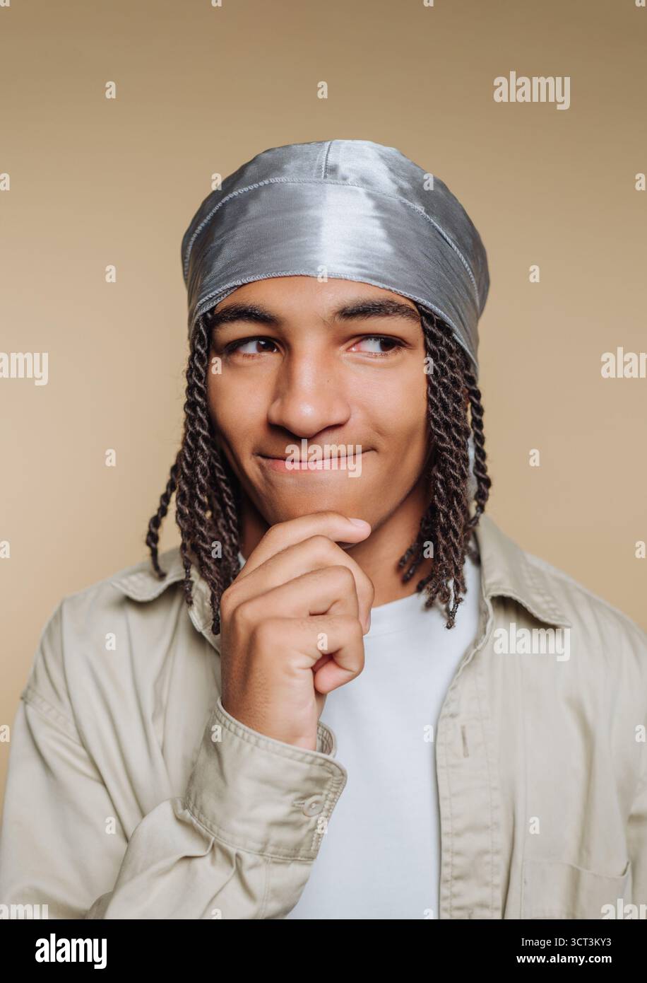 Un jeune homme avec des cheveux tressés pose avec soin, la main sur le menton, portant un enveloppement de tête argenté brillant devant un fond neutre. Son expression Banque D'Images
