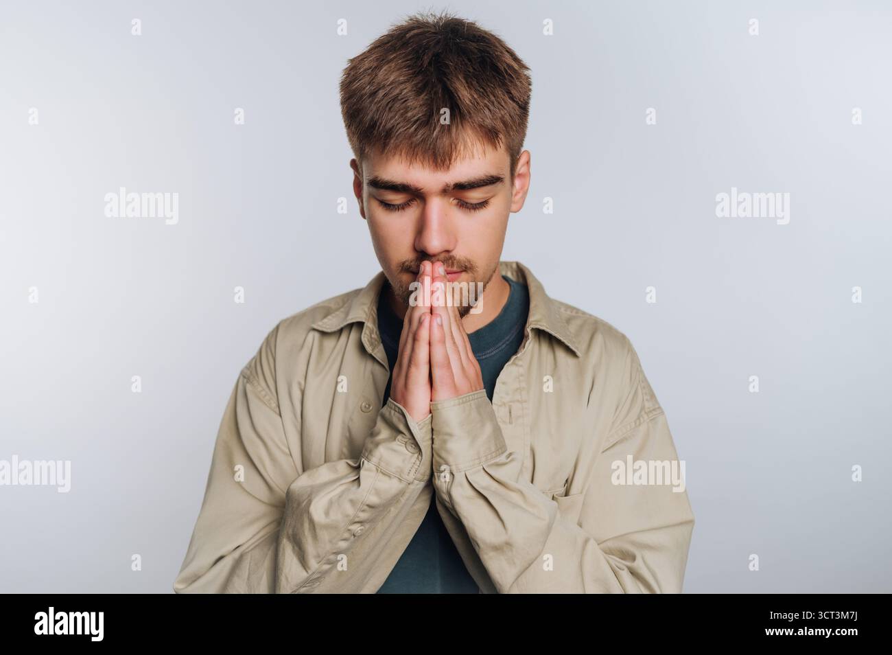 Un jeune homme se tient dans un espace intérieur bien éclairé, les yeux fermés et les mains pressées ensemble dans un geste de contemplation ou de prière. Il semble concentré et Banque D'Images