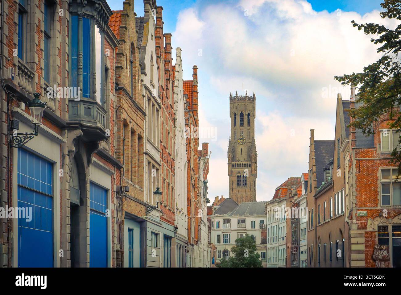 Une rue étroite à Bruges, en Belgique, avec des bâtiments traditionnels à pignon menant à la célèbre tour médiévale du beffroi contre un ciel nuageux. Banque D'Images