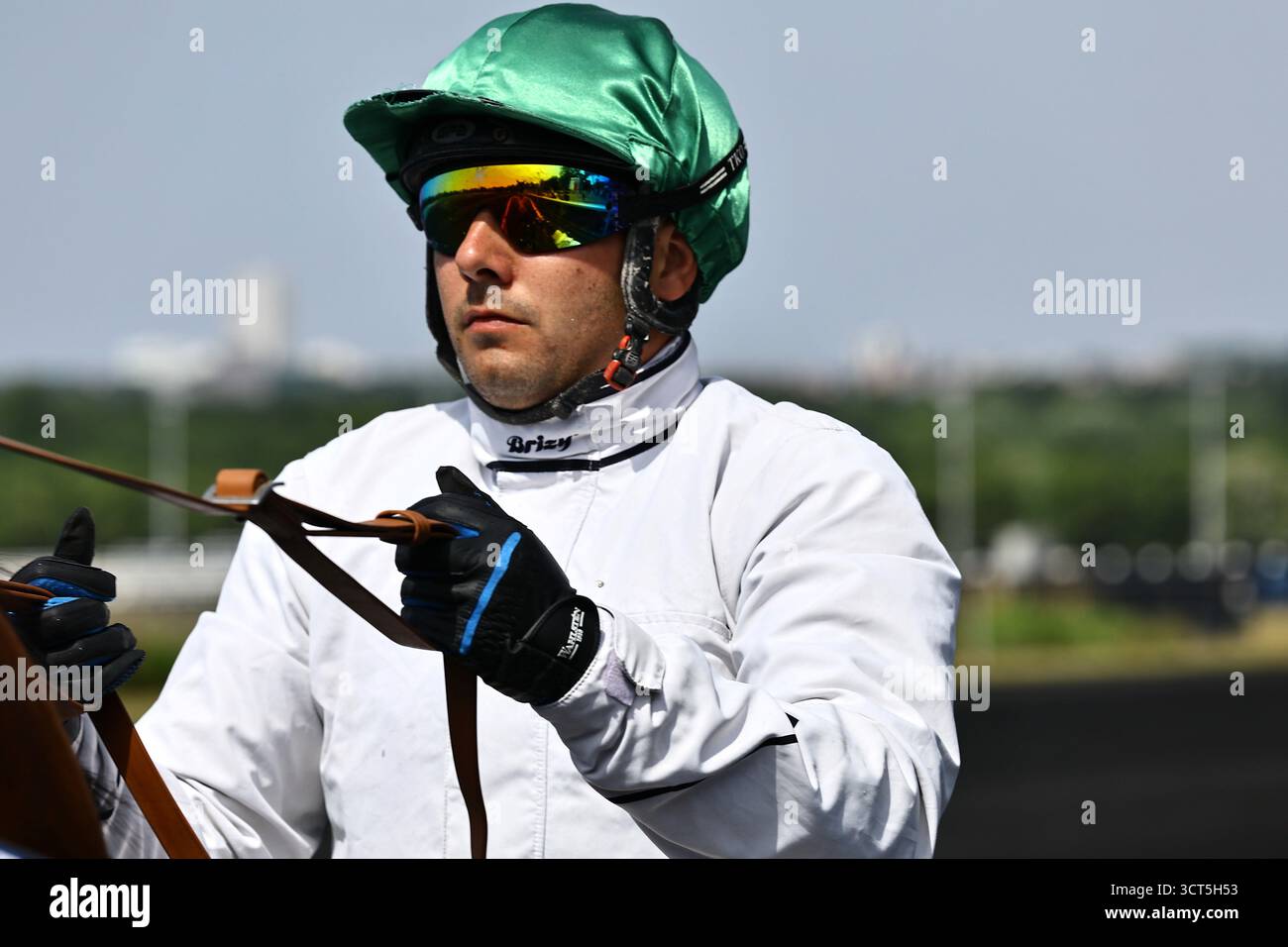 16.09.2025 Paris,Hippodrome de Vincennes.pilote de course harnais dans une veste blanche, casque vert, et lunettes de soleil miroirs tenant des rênes. Banque D'Images