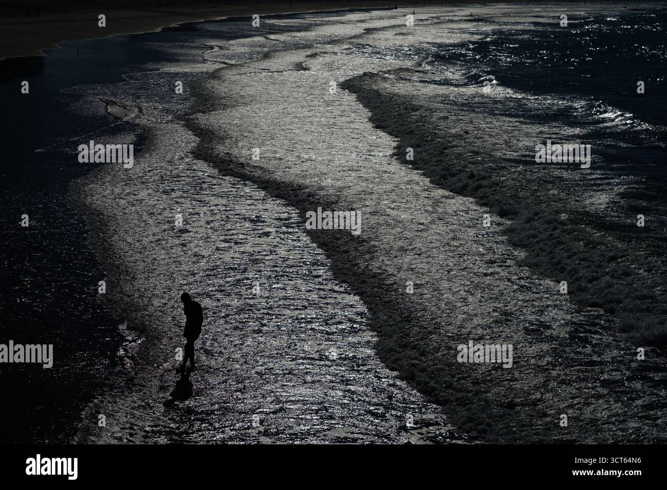Photographie discrète d'une silhouette d'une personne debout à la cheville dans les eaux peu profondes et brisant les vagues sur la plage. Banque D'Images