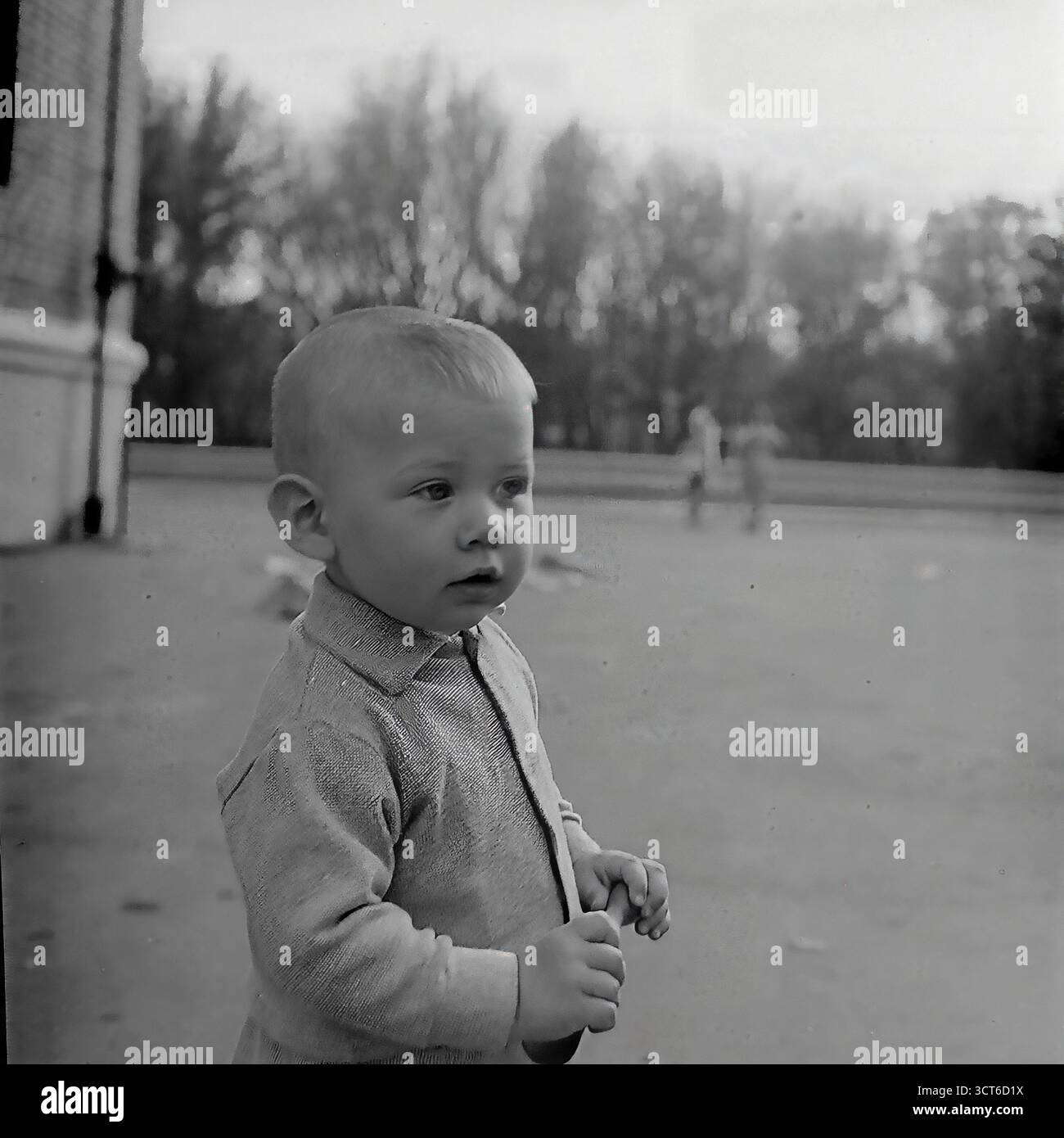 Portrait franc d'un jeune garçon dans un parc public ou une cour dans les années 1960 Sloviansk, RSS d'Ukraine. Cette photographie en noir et blanc capture l'expression innocente et contemplative de l'enfant. Il est vêtu d'un simple cardigan tricoté, typique de l'époque. En arrière-plan, les contours de craie pour un jeu de houssette sont visibles sur l'asphalte, plaçant la scène dans un contexte de jeux de rue quotidiens et de jeu en plein air. Cet instantané charmant et authentique d'un moment fugace évoque un puissant sentiment de nostalgie pour une enfance paisible de l'ère soviétique Banque D'Images