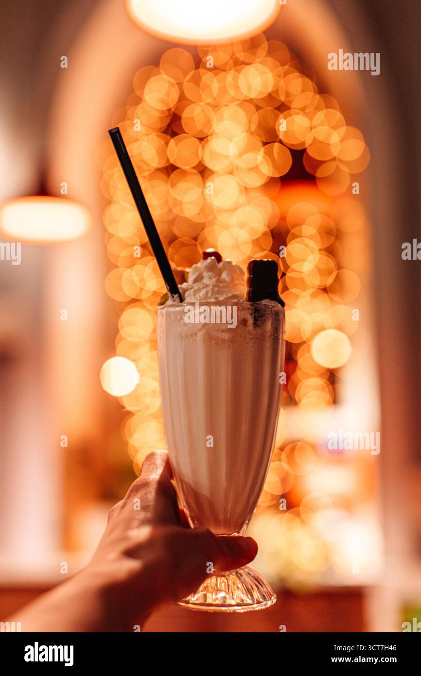Main de femme tenant un verre avec délicieux milkshake à la vanille avec des biscuits et de la crème fouettée sur fond de guirlande dorée Banque D'Images