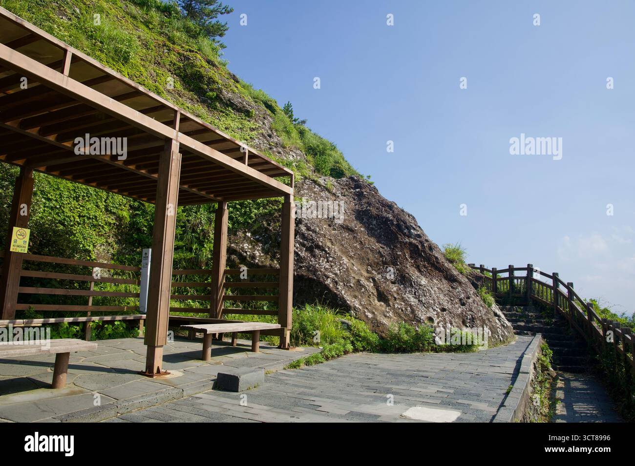 Un abri de repos en bois avec des bancs se trouve à côté du sentier pavé sur Seongsan Ilchulbong, un avis de non-fumeur sur son poteau, tandis que le sentier courbe autour d'un Banque D'Images
