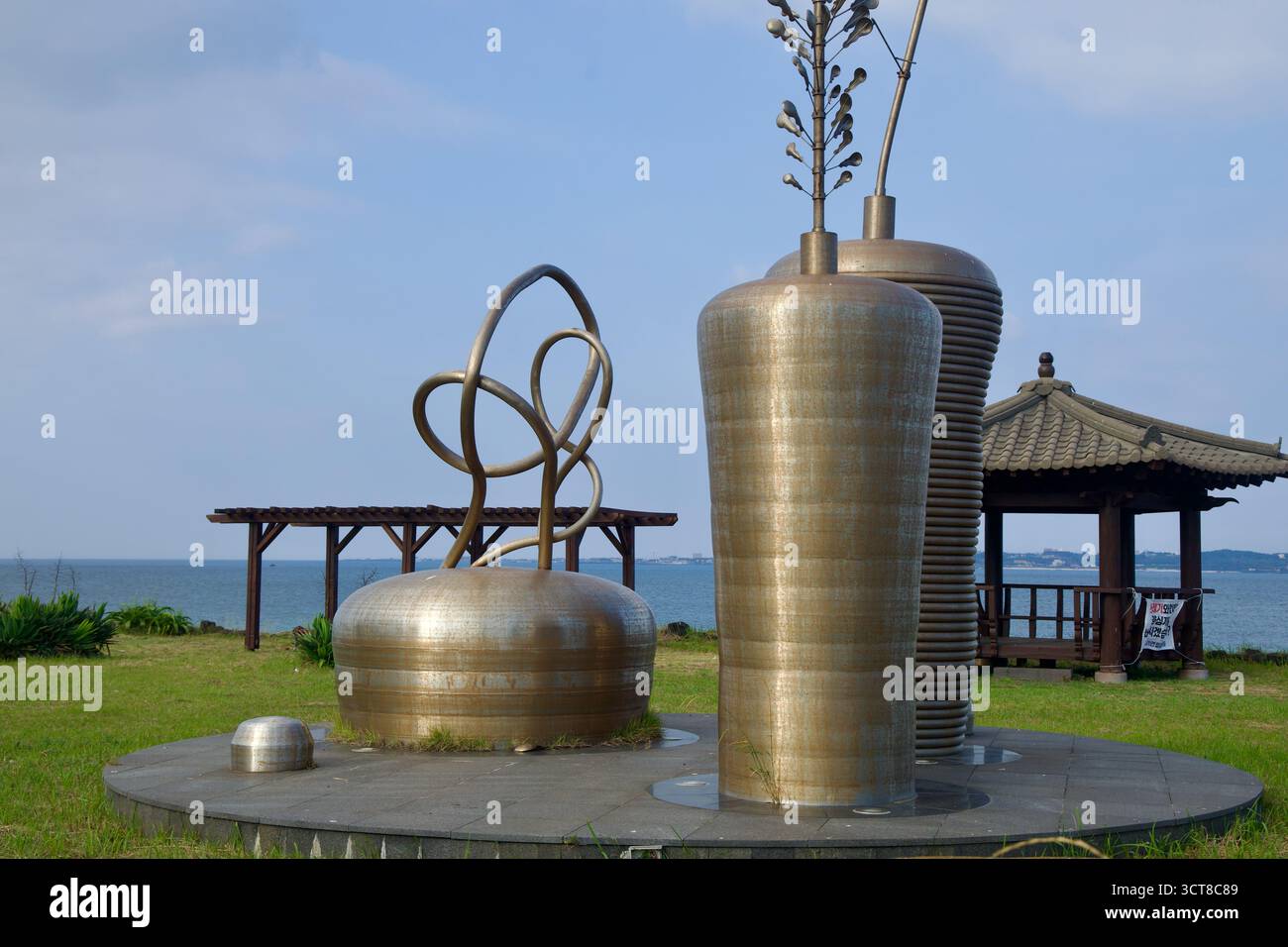 Une sculpture en métal poli se dresse sur la pelouse au bord de l'eau au port de Dumun à côté d'un pavillon en bois et de treillis, avec la mer ouverte qui s'étend au-delà de Jeju’ Banque D'Images
