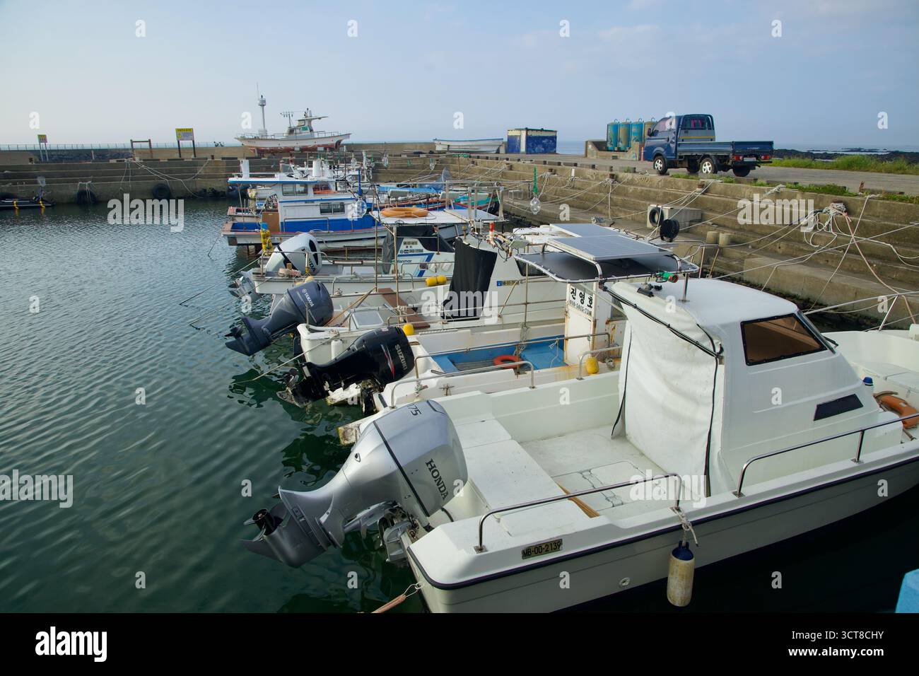 De petits bateaux de pêche équipés de moteurs hors-bord sont amarrés à l’intérieur du bassin en béton du port de Hado sur l’île de Jeju, avec des camions de travail et des engins portuaires le long du quai Banque D'Images