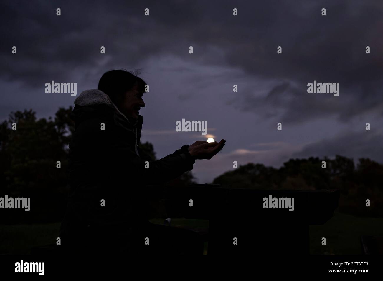 Silhouette en vue latérale d'une femme assise dehors dans l'obscurité, les mains en coupe comme si elle tenait la lune entre ses mains. Banque D'Images