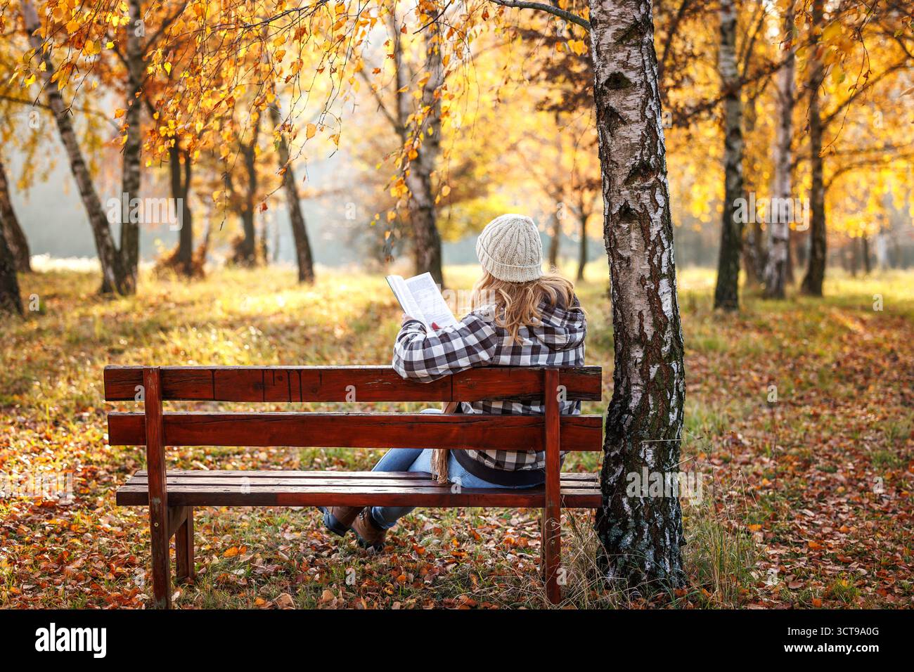 Femme se relaxant sur un banc en bois dans une forêt d'automne dorée, trouvant le bonheur et le bien-être mental tout en lisant un livre Banque D'Images