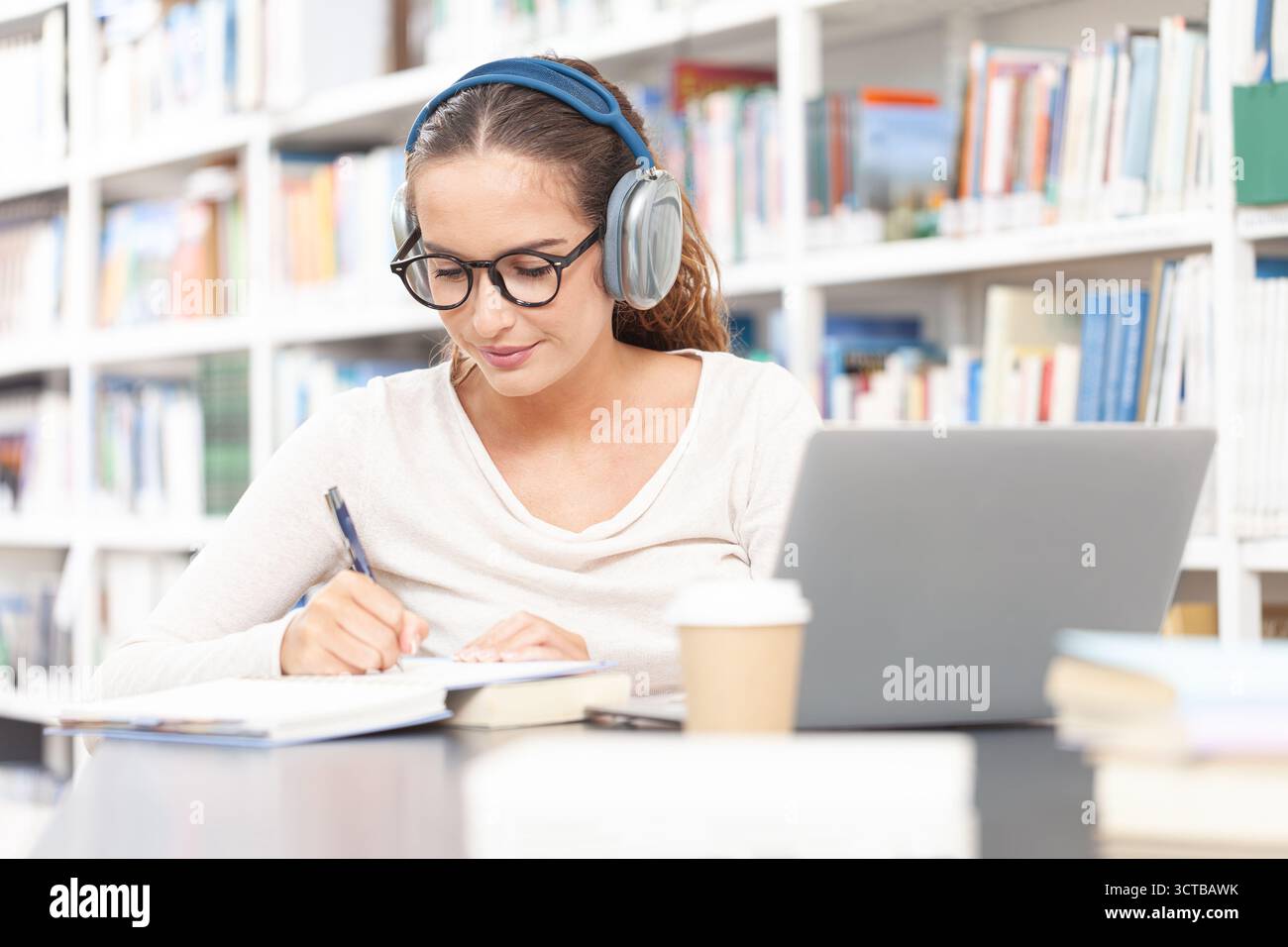 Jeune étudiante universitaire étudiante dans une bibliothèque, assise à un bureau avec un ordinateur, portant des écouteurs et prenant des notes dans un cahier. Concept o Banque D'Images