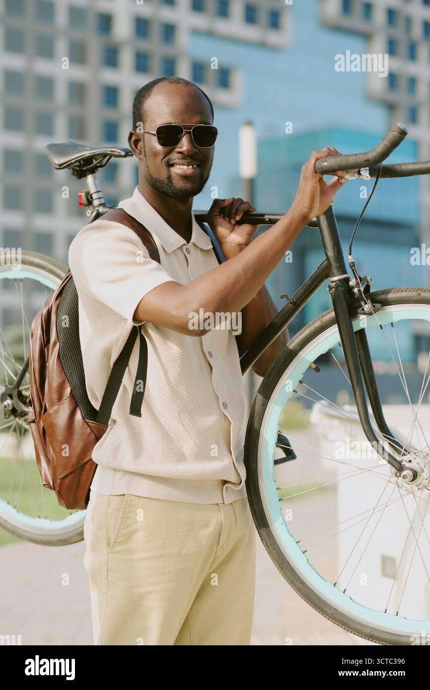 Jeune homme noir adulte souriant tout en transportant un vélo à l'extérieur dans la zone urbaine Banque D'Images