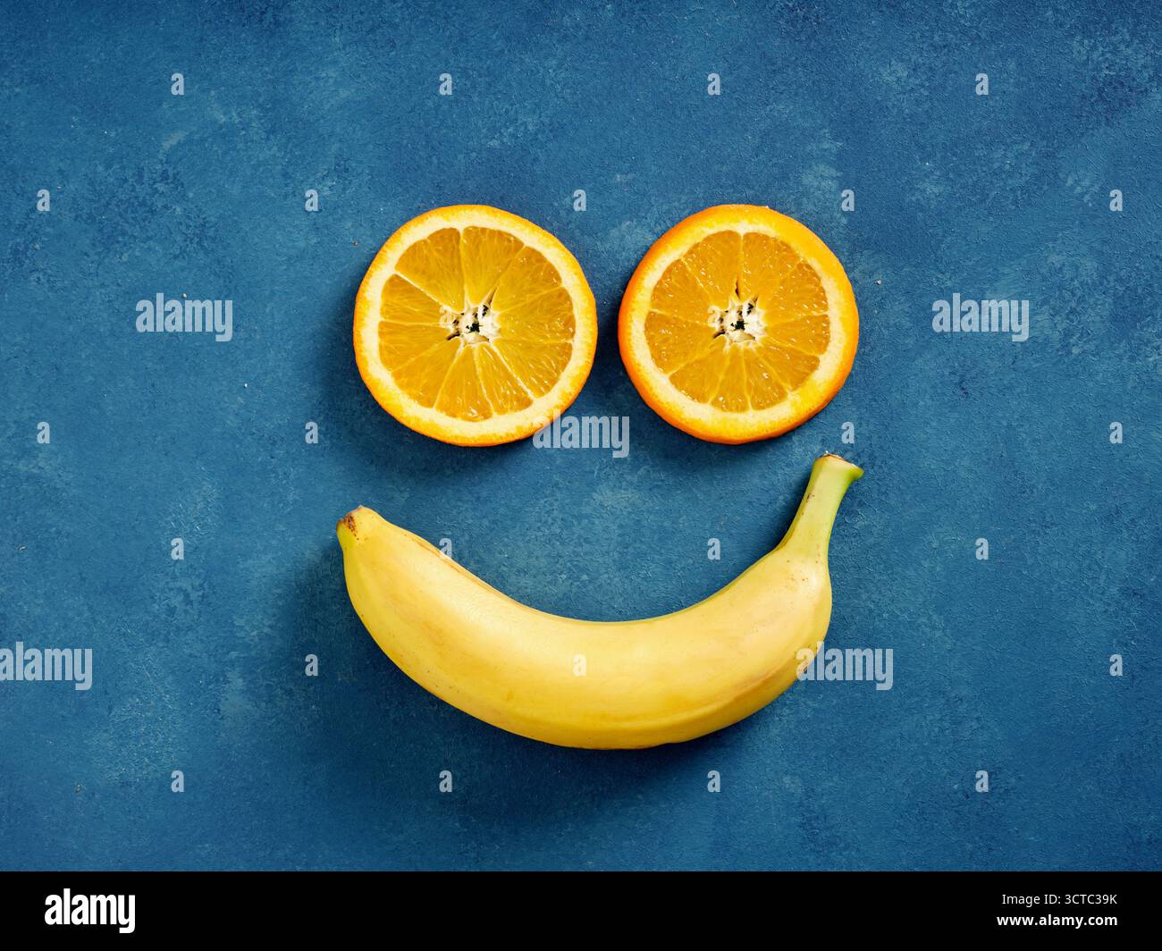 Tranches d'orange fraîches et une banane entière disposées comme un visage souriant joyeux sur un fond bleu, représentant le bonheur et une alimentation saine Banque D'Images