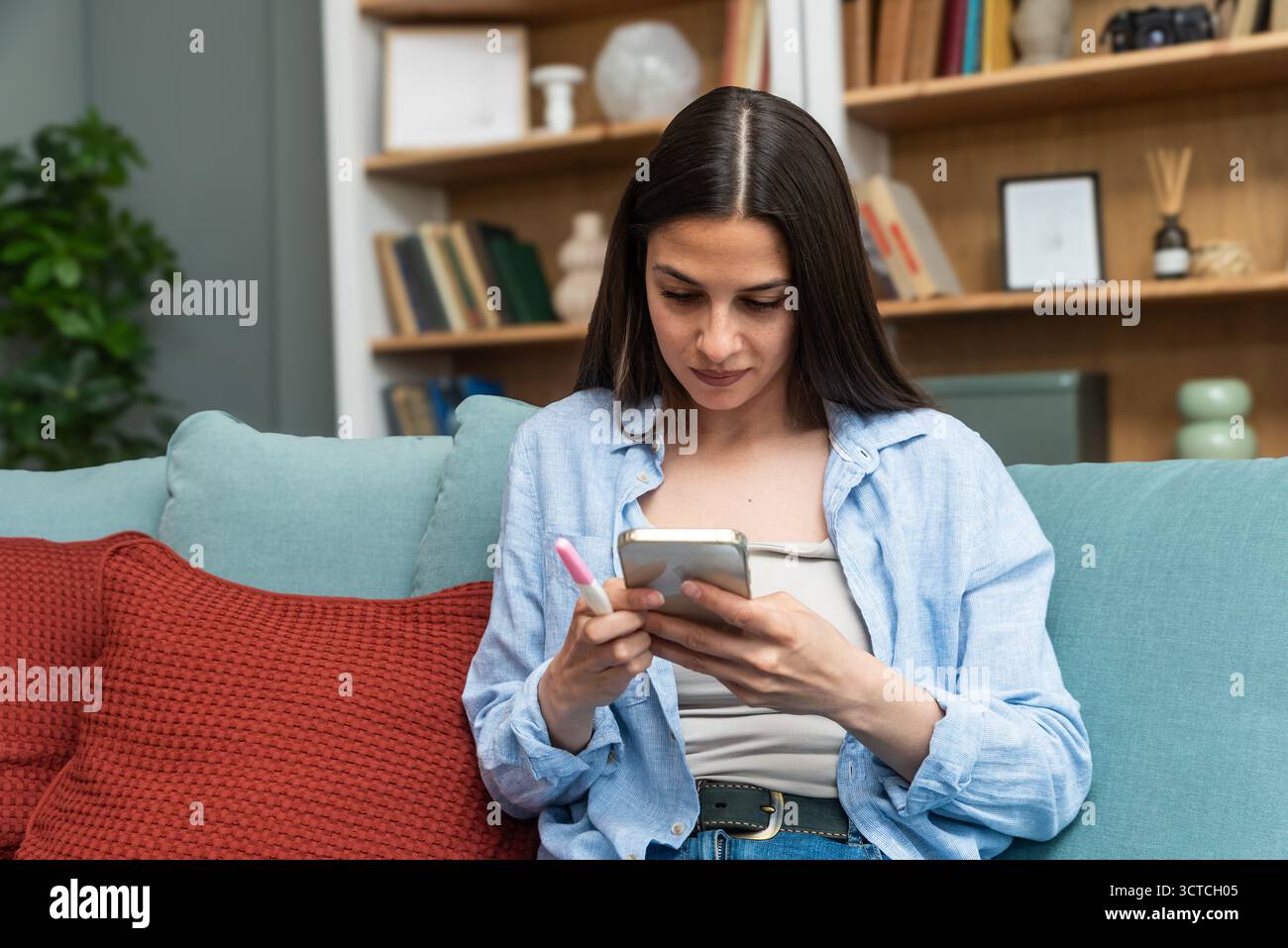 Jeune femme envoyant des SMS sur son smartphone tout en tenant un test de grossesse — en attendant les résultats, en envoyant des messages à un partenaire et en envisageant les prochaines étapes ; reproductif Banque D'Images