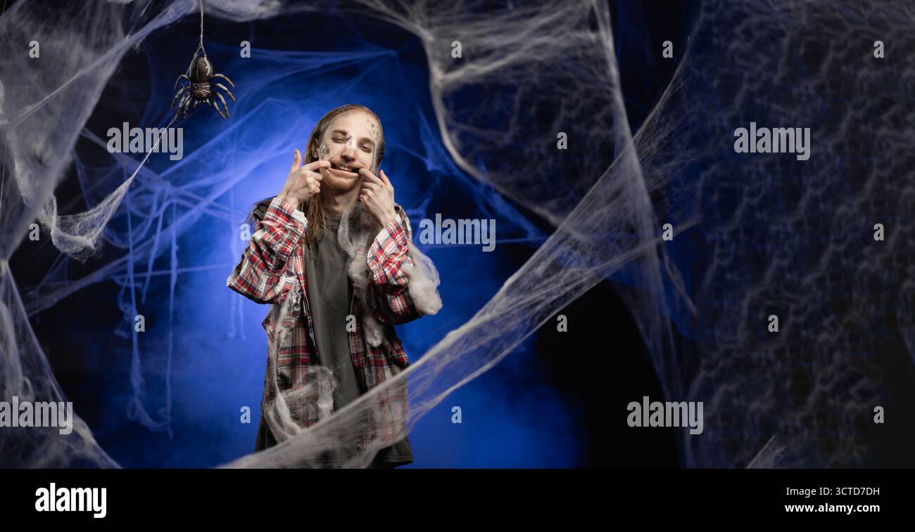 Mec mort avec horrible effrayant Halloween zombie maquillage sur le visage tient les doigts au bord des lèvres forces Smile a expression malheureuse debout couvert de spi Banque D'Images