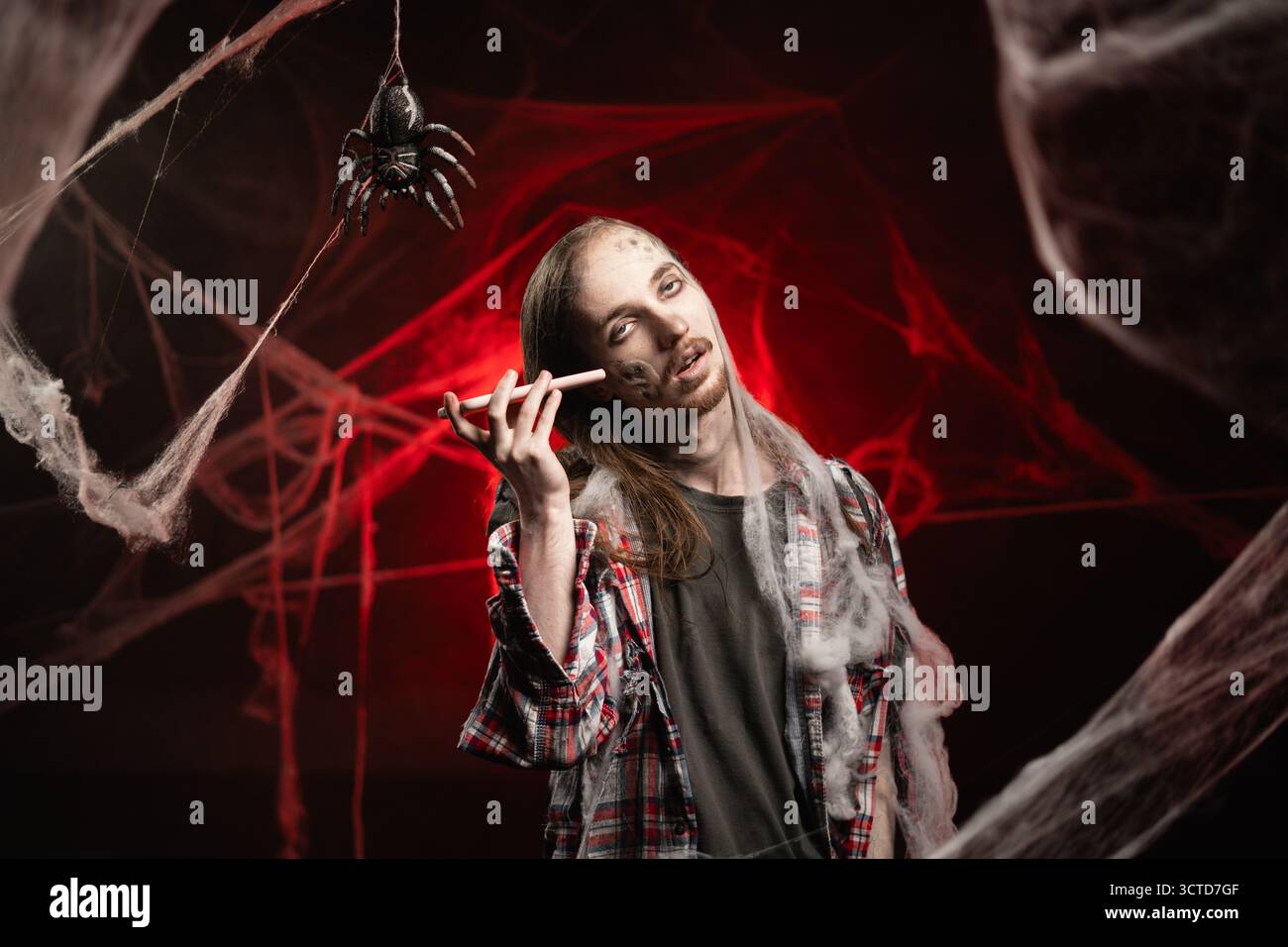 Homme effrayant avec le maquillage zombie effrayant d'Halloween piégé des toiles d'araignée écoutant un message vocal sur smartphone. Mec mort dans la toile de cob sur fond rouge sanglant. Banque D'Images