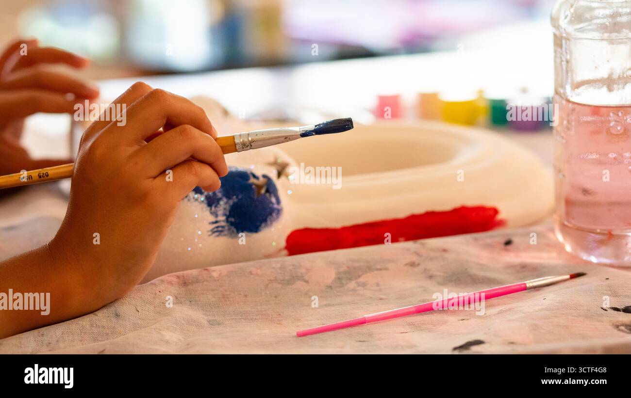 Main d’enfant tenant un pinceau tout en peignant un objet en céramique en bleu et rouge lors d’une activité artisanale. Projet de bricolage créatif avec des peintures Banque D'Images