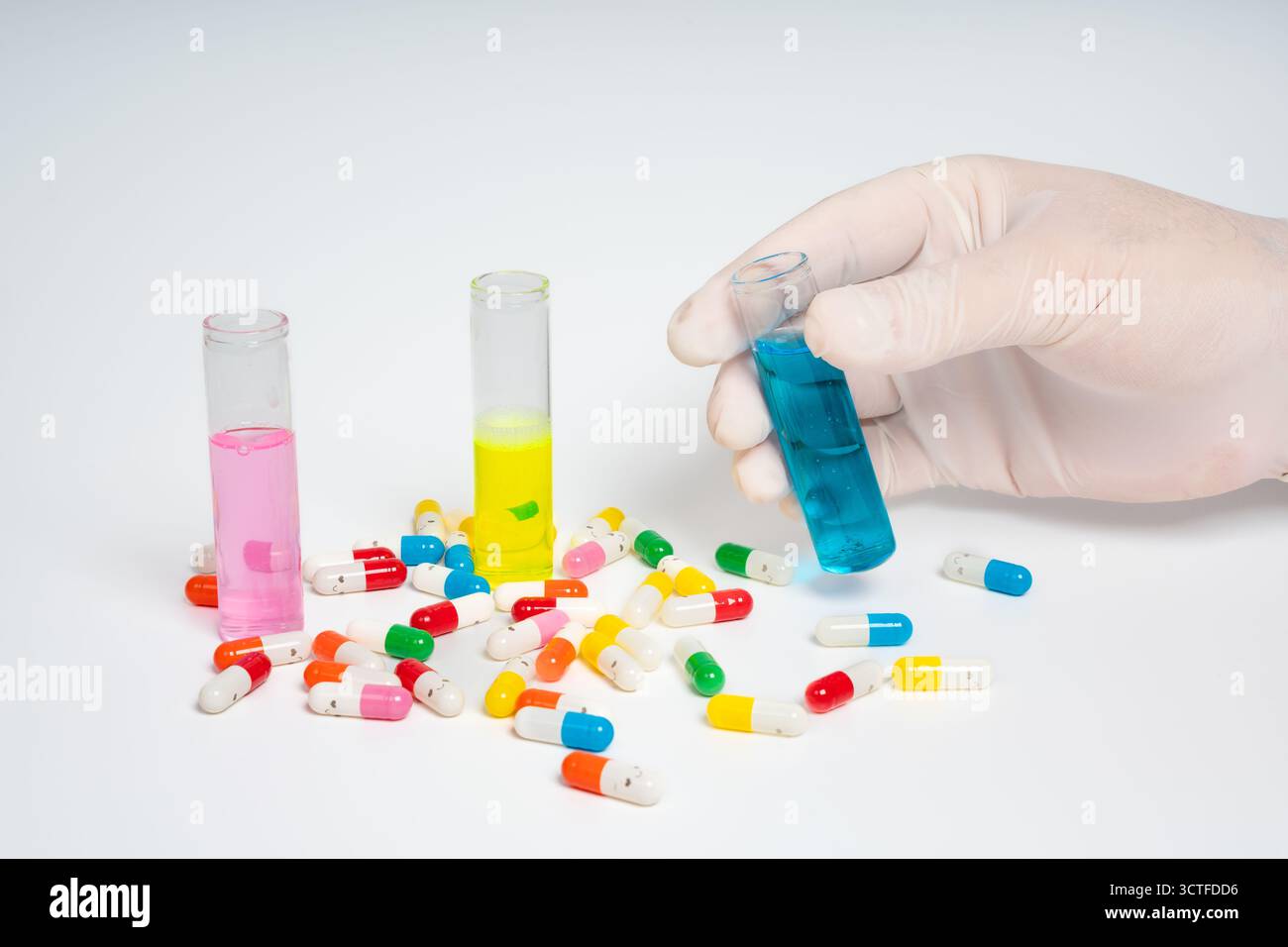 Main gantée tenant tube à essai avec liquide bleu, pilules colorées isolées sur fond blanc. Tubes à essai en verre et pilules tenus à la main Banque D'Images