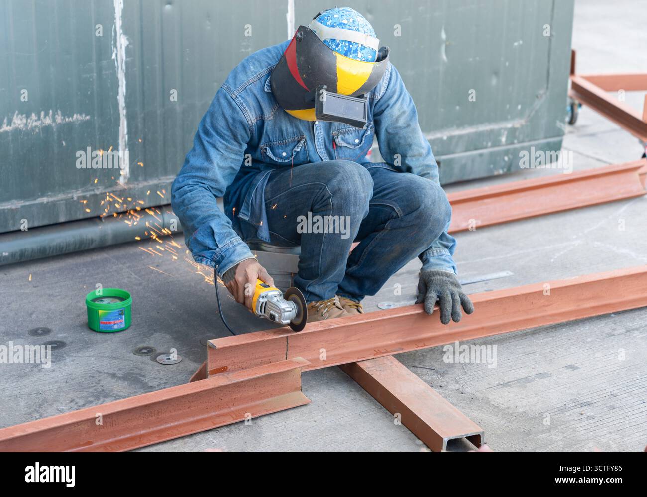 Un ouvrier de la construction portant un équipement de protection, y compris un casque de soudage et des gants, utilise une meuleuse d'angle pour couper une poutre en acier dans une industrie extérieure Banque D'Images