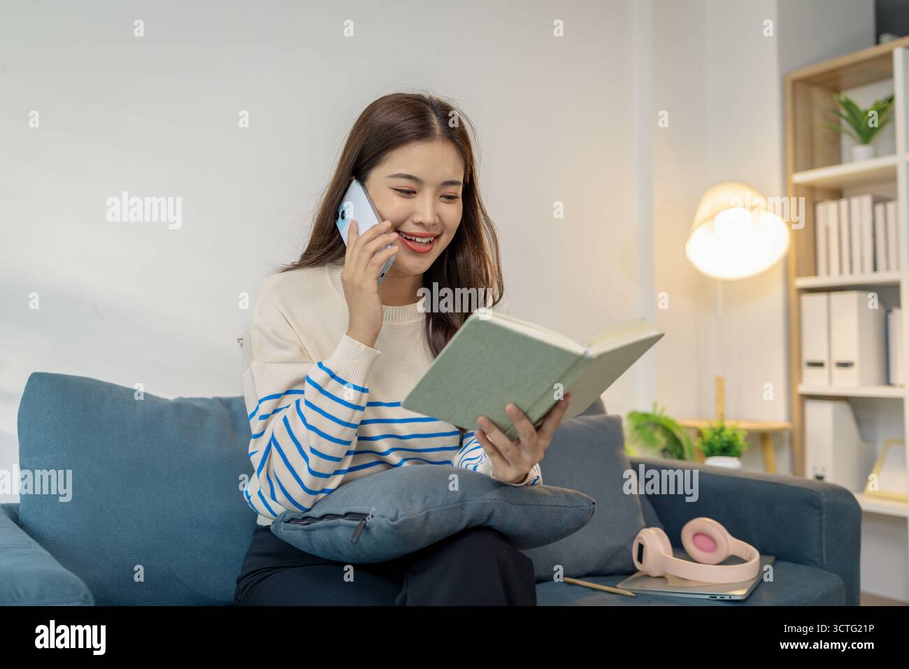 Une jeune femme assise confortablement sur un canapé, souriante tout en utilisant un smartphone avec un ordinateur portable sur ses genoux, montrant une scène de multitâche entre digi Banque D'Images