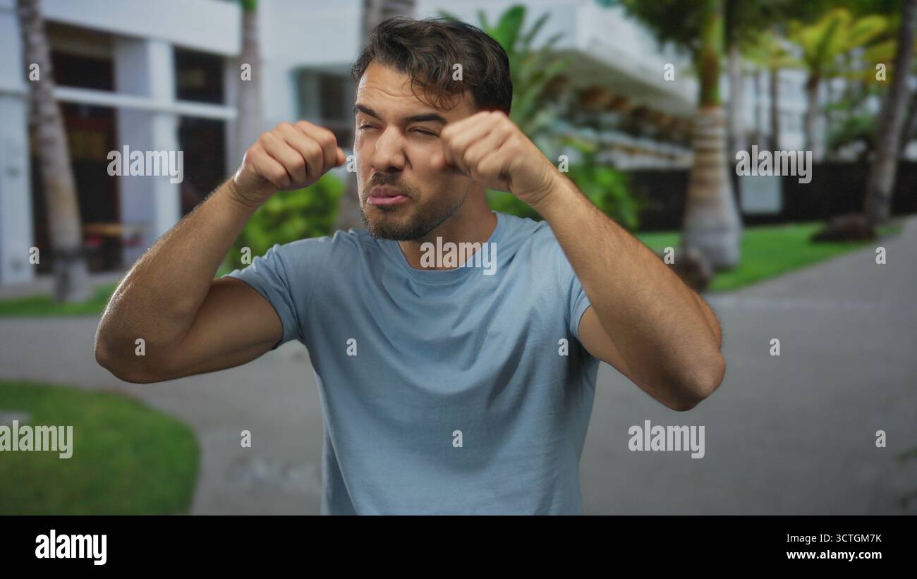 Un jeune homme hispanique à l'extérieur fait un geste de pleurs moqueurs dans un parc animé affichant une émotion et de l'humour ludiques sur fond de palmier Banque D'Images