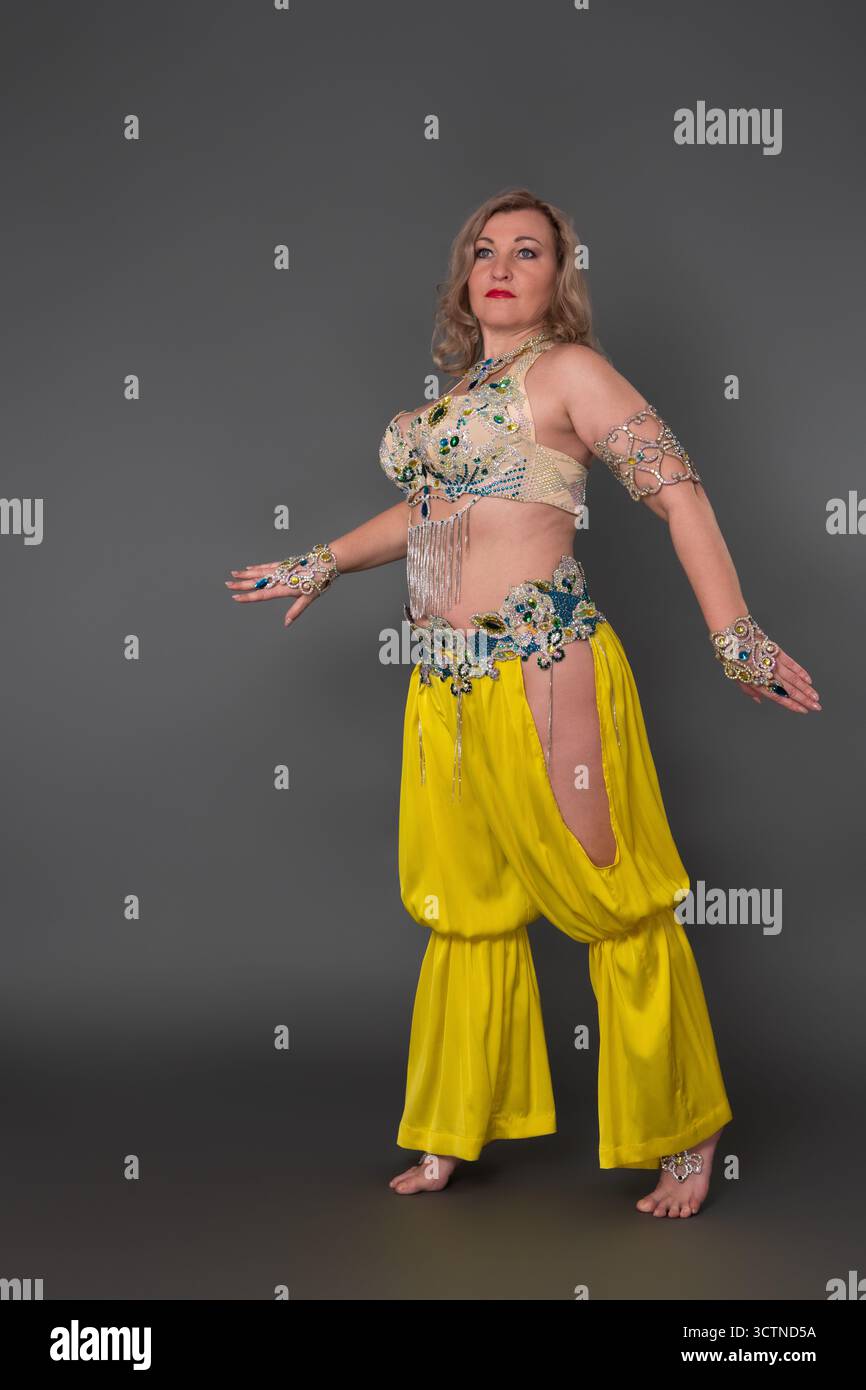 Danseuse sérieuse de la génération X enfilant en costume de danse - haut et pantalon de harem jaune se déplace pieds nus sur toute la longueur sur fond gris Banque D'Images