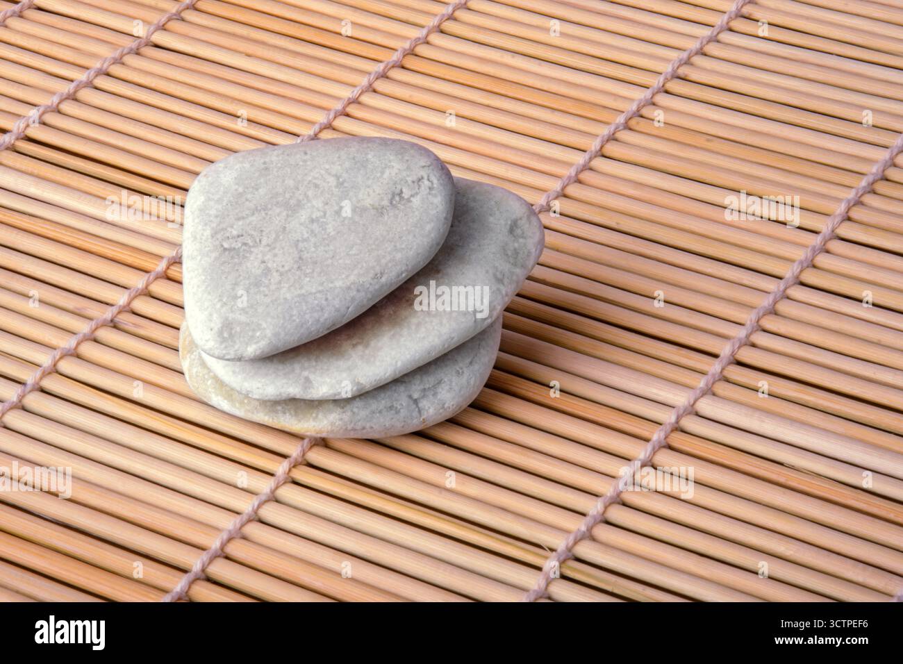 Une petite pile de trois galets gris lisses et plats disposés soigneusement sur un tapis de bambou. Le contraste entre les pierres fraîches et la texture chaude du bois. Banque D'Images