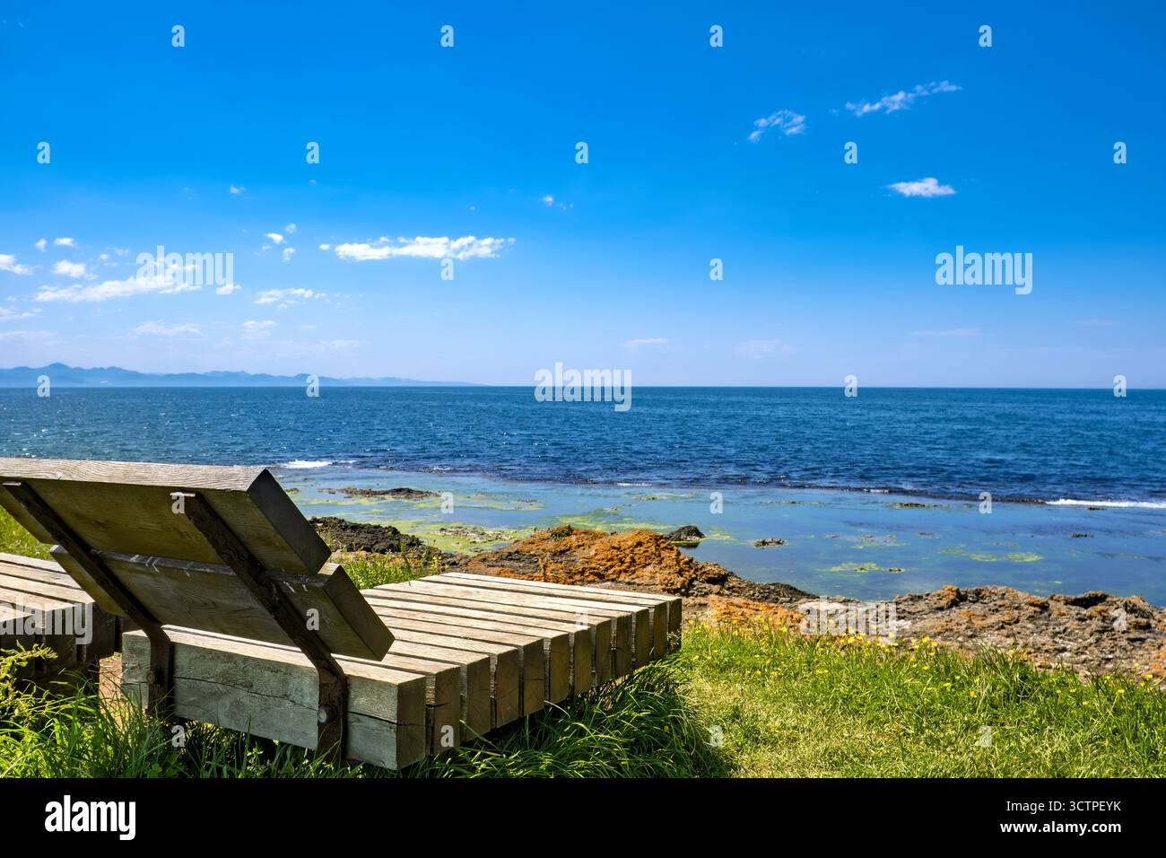 Un banc en bois se dresse sur des falaises verdoyantes en bord de mer surplombant la mer bleue. Le rivage rocheux et l'horizon créent une vue côtière paisible sous un ciel. Banque D'Images