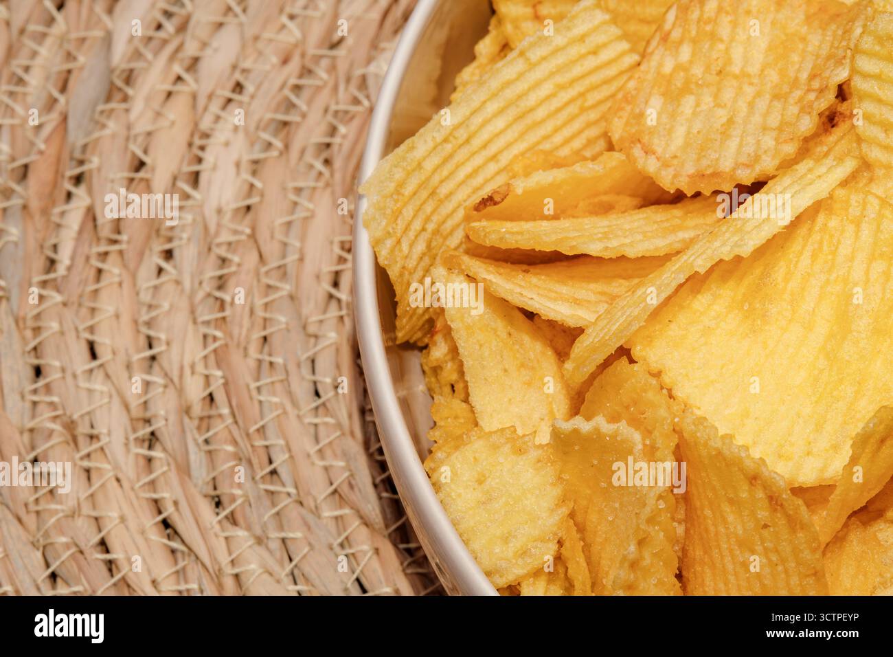 Gros plan d'un bol rempli de croustilles dorées croustillantes. La texture striée et le look croquant en font une collation salée classique. Placé sur un tapis naturel tissé Banque D'Images