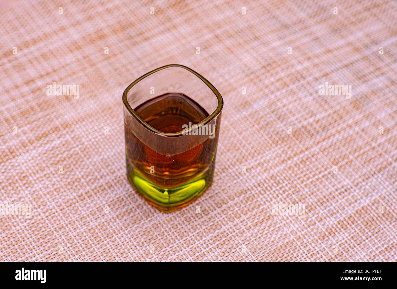 Isolé un verre à base teintée en vert, rempli d'une boisson ambre. Placé sur une surface beige texturée. La configuration simple et minimale. Banque D'Images
