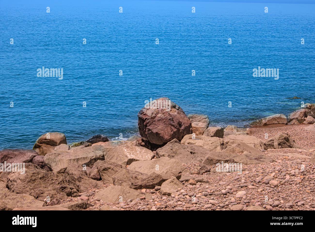 Rivage rocheux avec un grand rocher reposant près du bord de la mer bleue calme. Des cailloux et des pierres recouvrent la plage, se mélangeant à un horizon tranquille. Banque D'Images