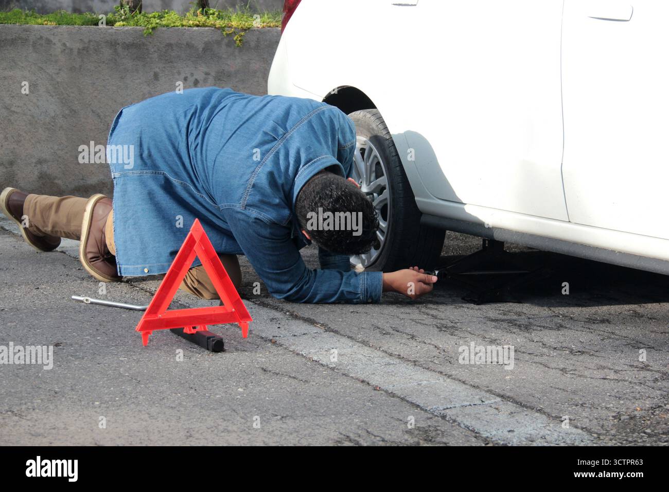 L'homme adulte latino à la peau foncée utilise un cric hydraulique pour changer le pneu crevé de sa voiture sur la route Banque D'Images
