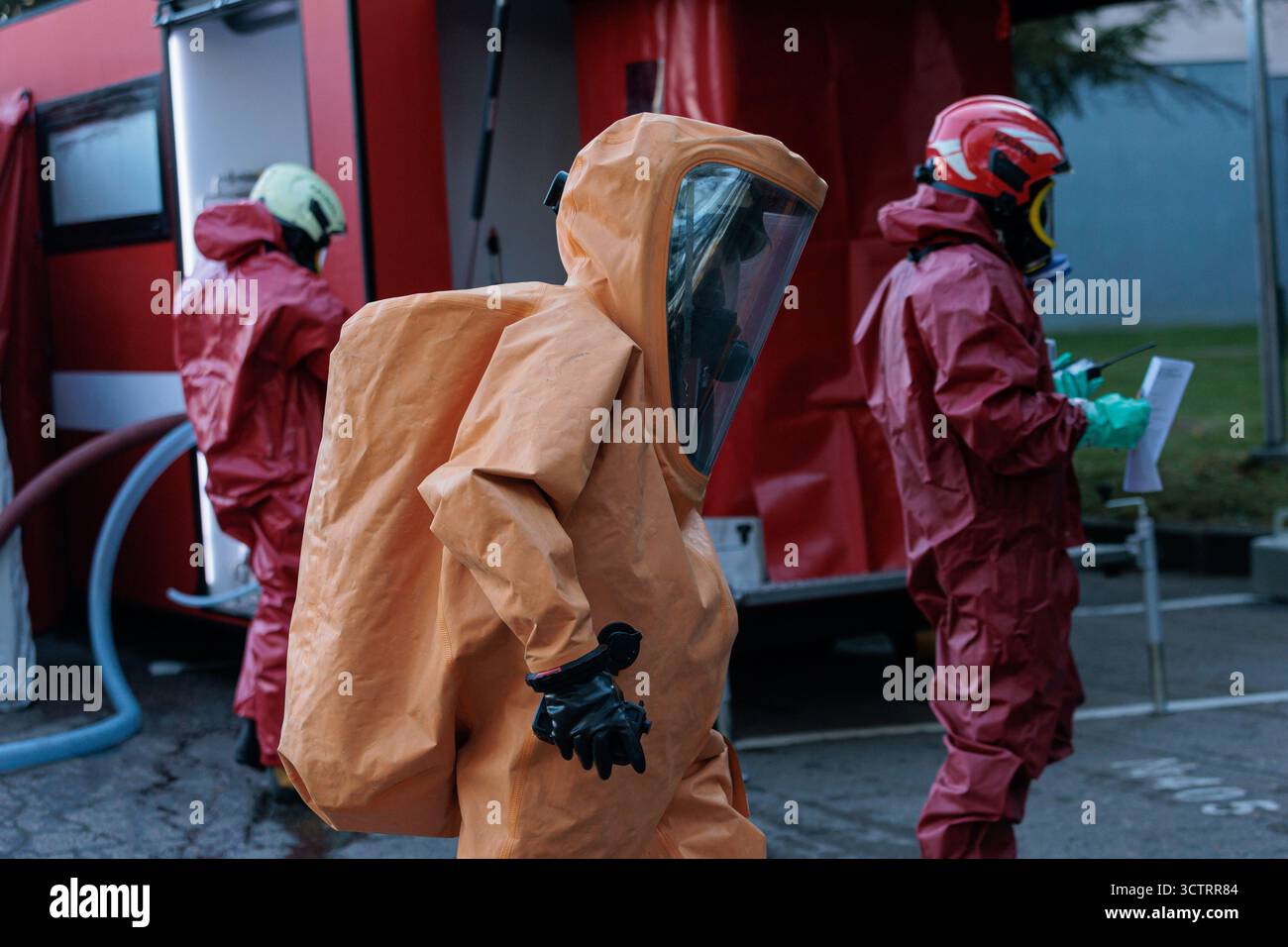 Les intervenants d'urgence vêtus de combinaisons de protection chimique orange et rouge et de masques intégraux se tenant à côté d'un véhicule d'intervention rouge lors d'une décontamination ou Banque D'Images