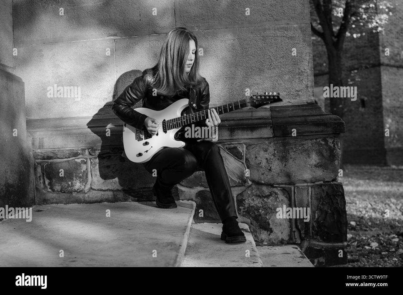 Fille guitariste en veste en cuir et pantalon en cuir avec guitare électrique blanche sur la rue de la ville Rock style Banque D'Images