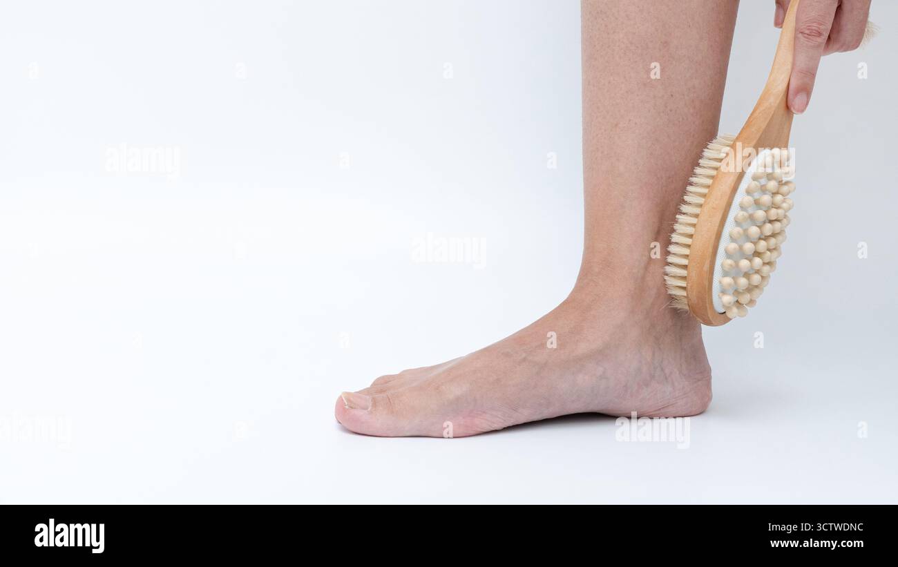 Une personne brosse doucement sa jambe avec une brosse corporelle en bois, mettant l'accent sur un rituel apaisant d'auto-soin dans un fond propre et minimal Banque D'Images