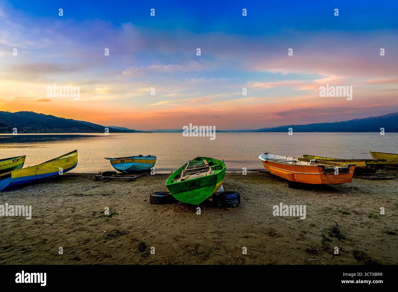 Bateaux de pêche colorés reposant sur la rive sablonneuse du lac Ohrid au coucher du soleil, avec une eau calme et des montagnes lointaines à Pogradec, Albanie Banque D'Images