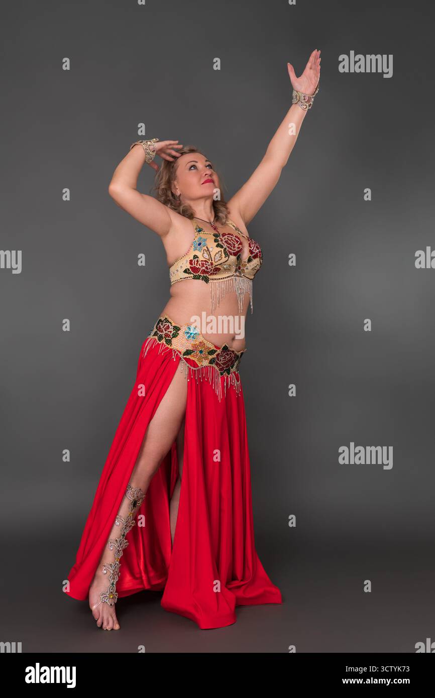 Femme adulte mature danseuse avec les bras levés vêtu d'une tenue de danse - haut de soutien-gorge et jupe longue rouge avec fente se déplaçant pieds nus sur toute la longueur contre le gris Banque D'Images