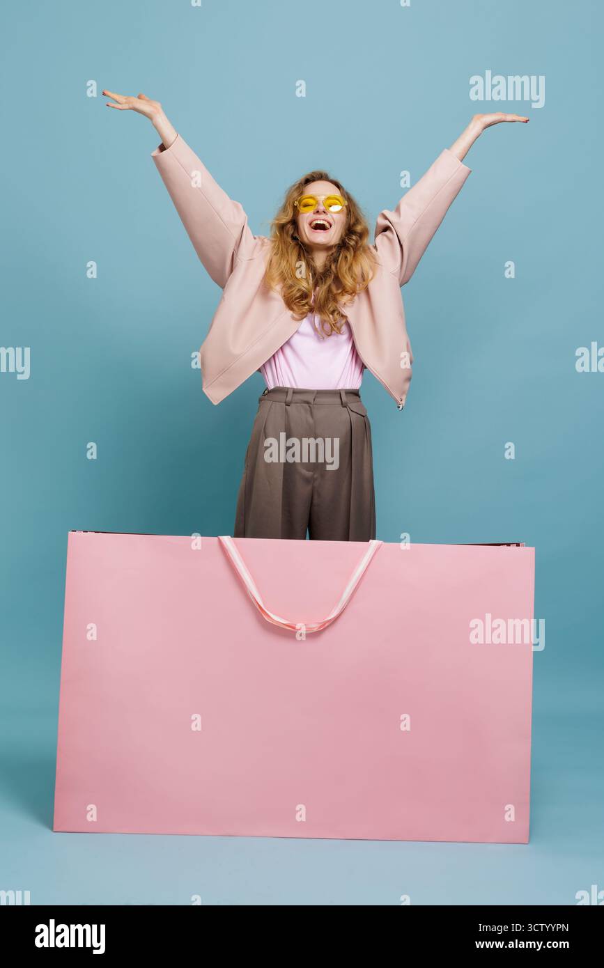Acheteur excité levant les bras derrière un sac rose surdimensionné sur fond bleu, parfait pour le Black Friday, les remises, les publicités au détail, les bannières et les campagnes sociales Banque D'Images
