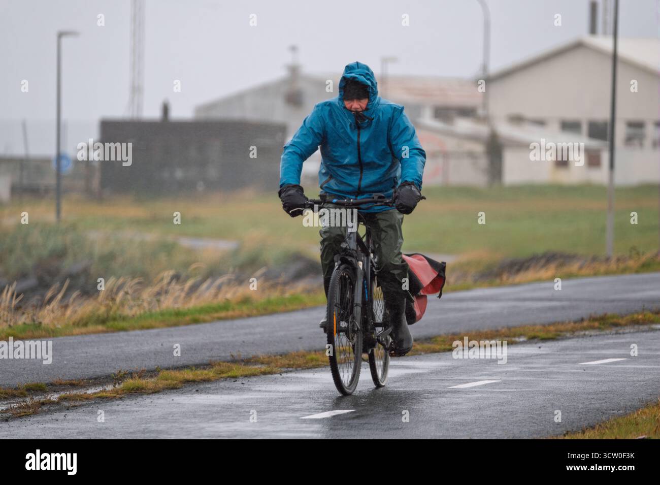Cycliste luttant contre la tempête et la pluie sur une route islandaise. Pulvérisation, rafales et faible luminosité ; gravillons et résilience par temps brutal. Banque D'Images