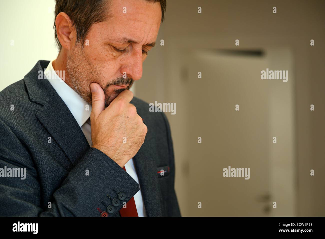 Homme d'affaires inquiet en costume élégant pensant avec la main sur le menton, debout dans l'intérieur du bureau. Mise au point sélective. Banque D'Images