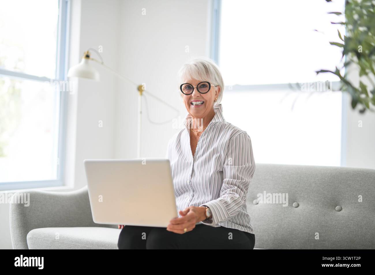 Un Portrait de femme âgée sur fond gris assis sur un canapé avec ordinateur portable Banque D'Images