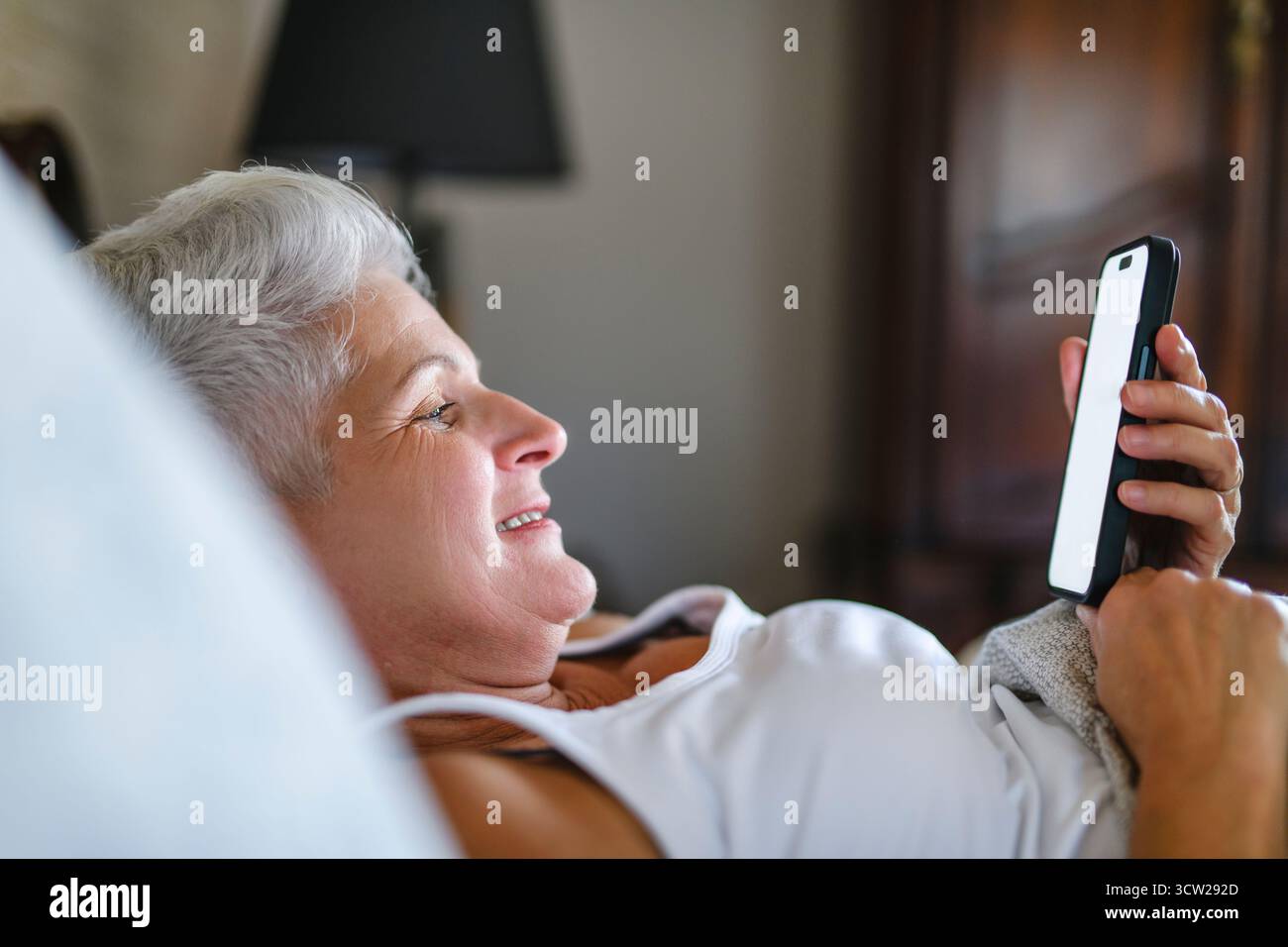 Une femme aînée à la maison couchée sur le lit sous couette à la chambre à coucher en utilisant un téléphone portable Banque D'Images