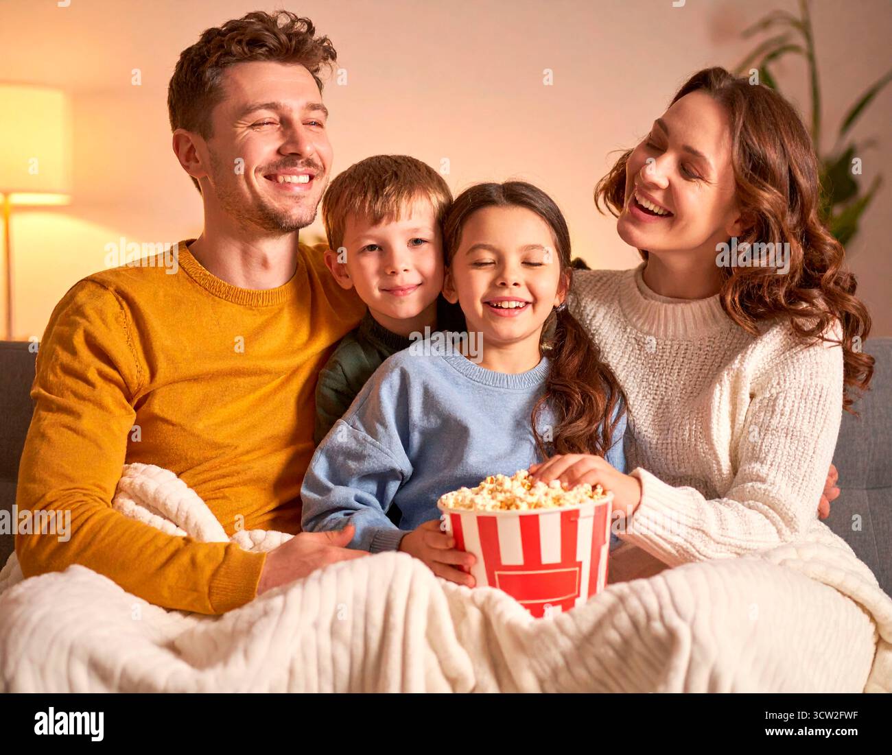 Famille joyeuse riant et mangeant du pop-corn assis sur le canapé sous une couverture Banque D'Images