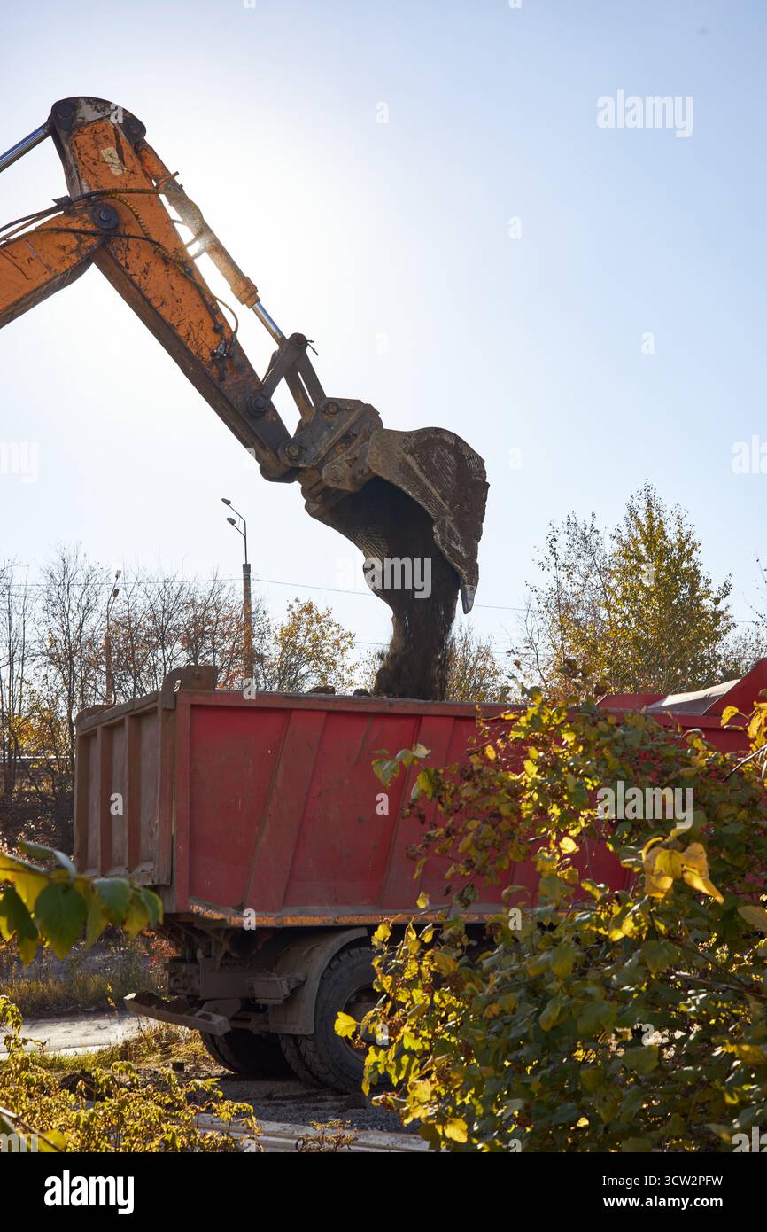excavatrice verse du béton dans un camion rouge sous un ciel bleu clair sur un chantier de construction. Les arbres d'automne entourent la région, ajoutant à l'atmosp saisonnière Banque D'Images