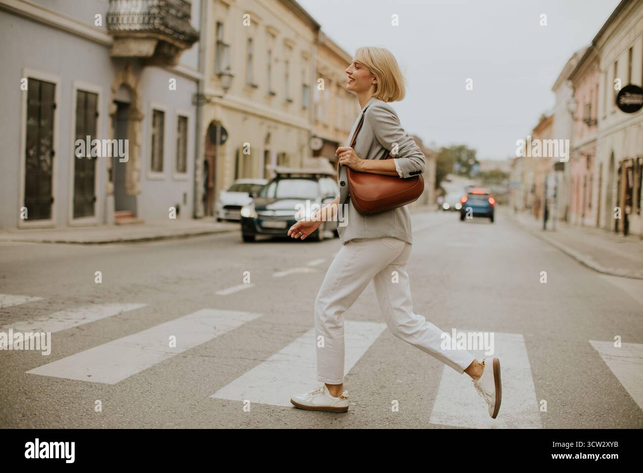 Une femme marche en toute confiance à travers une traversée de zèbre dans une ville. Elle porte un blazer gris clair, un pantalon blanc et des baskets. Les véhicules sont garés à proximité comme s Banque D'Images