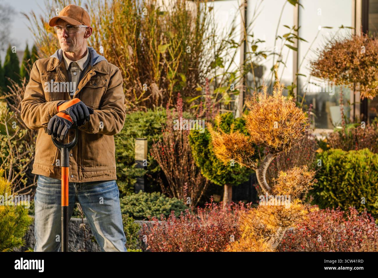 Un homme se tient debout avec une pelle dans une pépinière de plantes, entouré de diverses plantes et arbustes, profitant de la lumière chaude de l'après-midi tout en se préparant au jardinin Banque D'Images