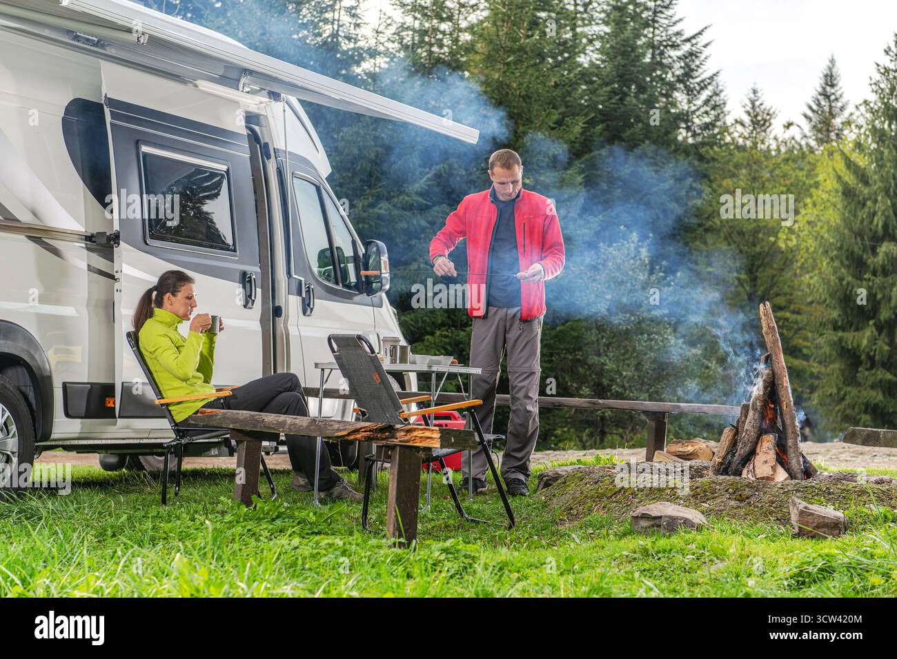 Un homme prépare de la nourriture sur un feu de camp tandis qu'une femme profite d'une boisson chaude assise près d'un camping-car dans un cadre forestier serein. Banque D'Images