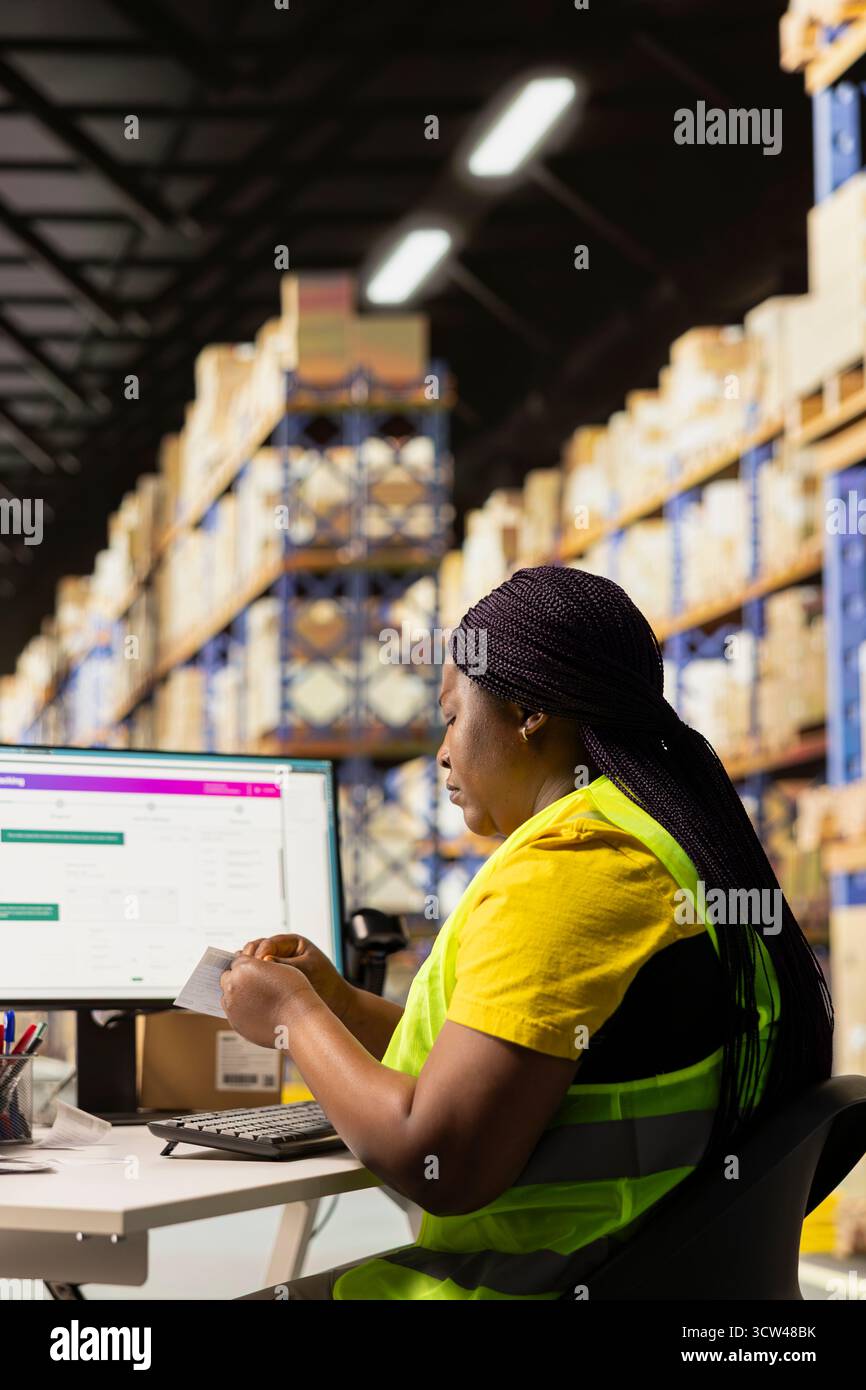 Employé noir de la réserve traitant les étiquettes de facture de transport aérien pour les marchandises, assurant des détails d'expédition précis sur le logiciel de suivi des colis. Femme en gilet hi vis au travail industriel. Banque D'Images