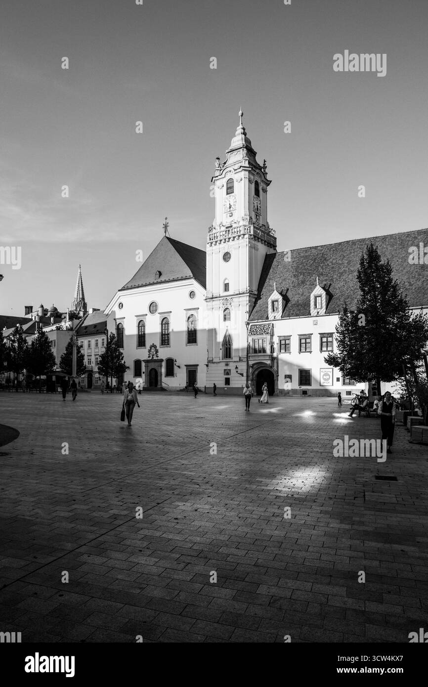Les gens se promènent sur une grande place historique avec une tour de l'horloge proéminente. Le soleil projette une lumière chaude sur les bâtiments et les arbres environnants. Banque D'Images