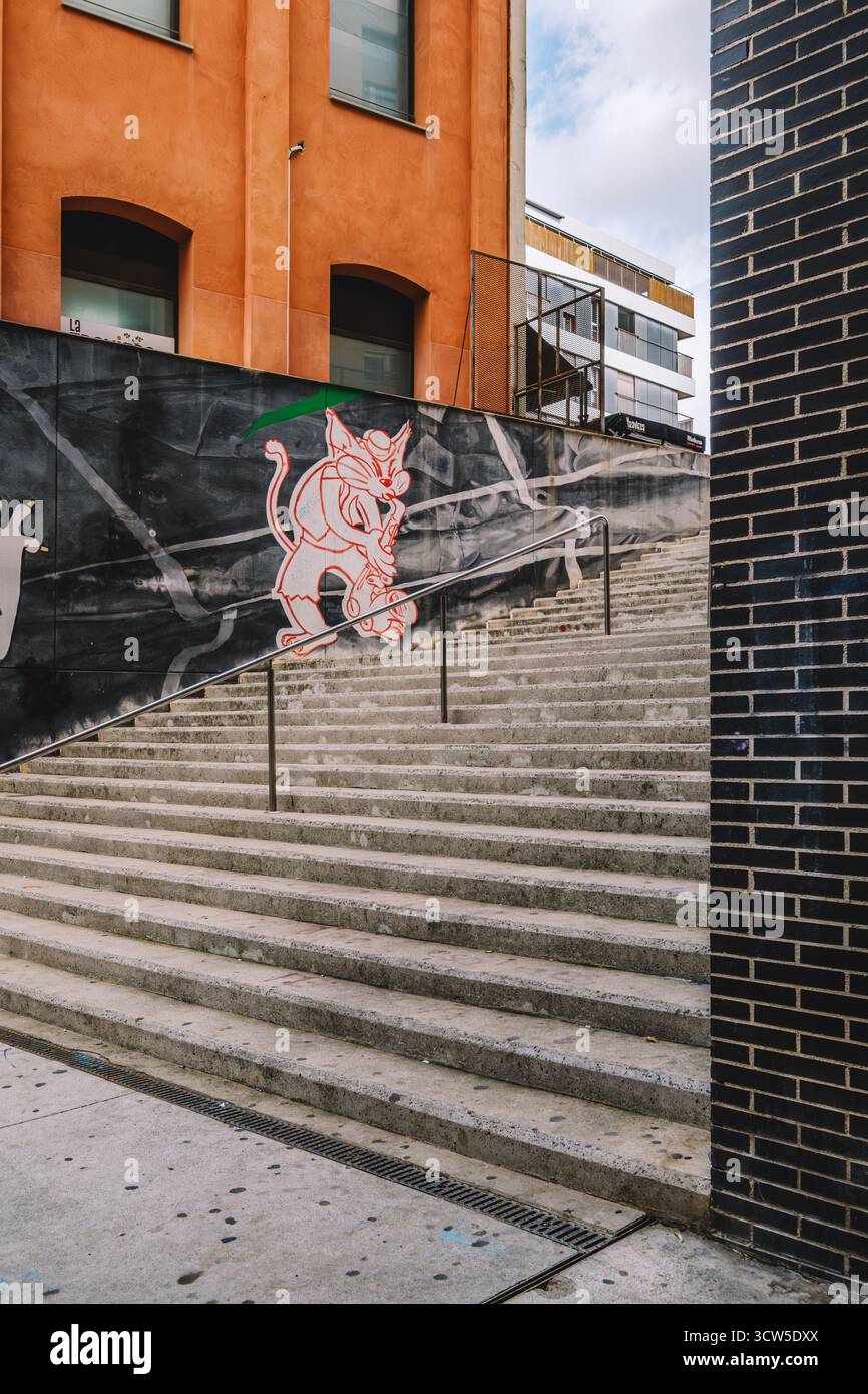 Vue sur un escalier extérieur en béton flanqué d'un bâtiment orange et d'un mur de briques sombres, orné d'une fresque murale expressive représentant un chat. Idéal pour urba Banque D'Images