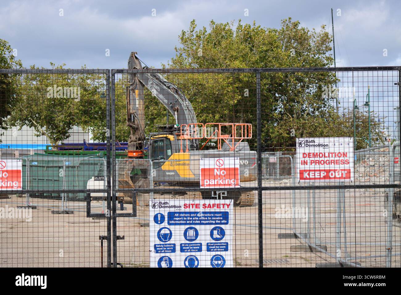 Démolition en cours de l'ancienne gare routière de Gosport, Hampshire, Angleterre. Un site de démolition entouré de clôtures métalliques dispose d'une grande pelle hydraulique en fonctionnement actif. Une signalisation bien visible avertit que « DÉMOLITION EN COURS, NE PAS ENTRER », ainsi que « pas de stationnement » et des instructions de sécurité détaillées. Les arbres et les barrières de construction encadrent l'arrière-plan, soulignant la tension entre la perturbation industrielle et la sécurité des procédures. Banque D'Images