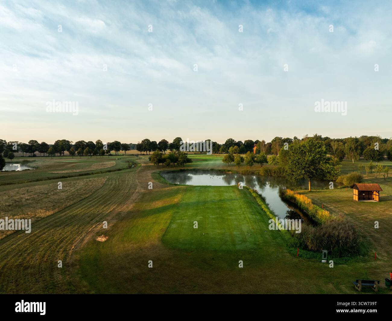 Vue drone d'un parcours de golf avec fairway vert et danger d'eau dans la lumière chaude du matin. L'image montre des arbres, des reflets sur l'étang et du ski clair Banque D'Images